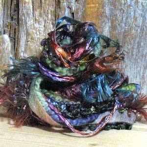 May include: A colorful, textured art piece composed of various yarns and ribbons in shades of purple, green, blue, and red. The piece has a layered, organic form, with fringed and looped elements.