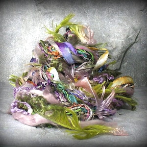 May include: A pile of colorful craft yarn in shades of green, purple, pink, and gold. The yarn appears to be a mix of textures, including ribbon, fringe, and fluffy fibers. The overall effect is a vibrant and textured display, suitable for various crafting projects.