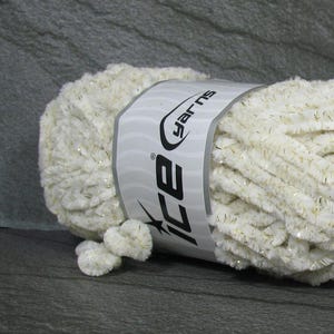 May include: A skein of soft, cream-colored chenille yarn featuring subtle gold metallic flecks throughout the fibers. A white paper band wraps around the center of the yarn, displaying the brand name Ice Yarns in black text with a logo.