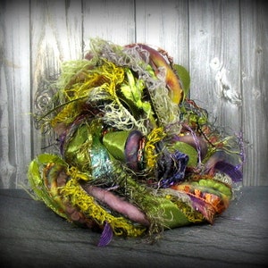 May include: A textured, multi-colored fiber art piece. The artwork features a mix of green, yellow, purple, and orange fibers, ribbons, and yarns. The piece has a layered, organic appearance, with a variety of textures and materials.