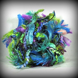 May include: A ball of yarn made of various shades of blue, green, and purple yarn with some metallic threads. The yarn is arranged in a loose, fluffy ball.