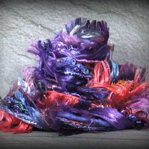 May include: A pile of colorful, textured fabric in shades of purple, red, and blue. The fabric appears to be layered and ruffled, with a shimmering, iridescent quality. The overall effect is rich and decorative.