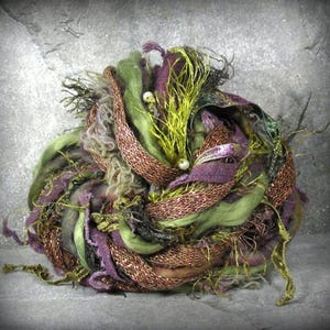 May include: A ball of mixed fiber art yarn in shades of green, purple, and bronze. The yarn includes various textures, such as woven ribbon, fuzzy strands, and metallic threads. Small white beads are interspersed throughout the yarn ball.