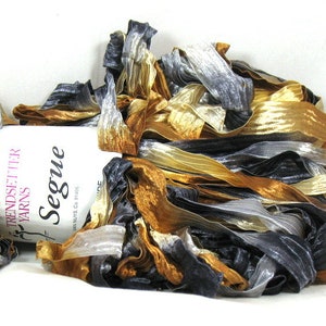 May include: A skein of Trendsetter Yarns Segue ribbon yarn featuring a metallic color palette of gold, silver, and black. The flat, textured ribbon strands are wrapped around a white paper label with the text Trendsetter Yarns Segue.