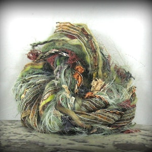 May include: A ball of mixed fiber yarn in shades of green, brown, and gold. The yarn has a textured appearance with various threads and ribbons woven together. The color palette includes olive, rust, and metallic accents, creating a unique craft supply.