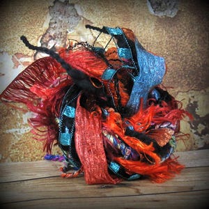 May include: A colorful yarn ball made of various textures and colors, including red, orange, black, blue, and green. The yarn is arranged in a loose, organic shape, creating a visually interesting and tactile piece.