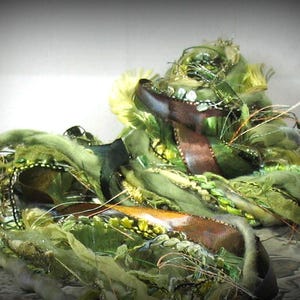 May include: An abstract art piece featuring a layered composition of green, brown, and gold textures. The artwork incorporates ribbons, fabric, and decorative elements, creating a unique and textured appearance. The piece has a handmade, organic feel.