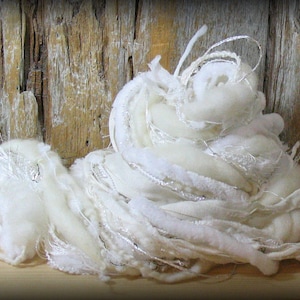 May include: A close-up of a pile of white yarn with various textures. The yarn includes fluffy wool, woven ribbons, and strands of silver thread. The background is a weathered wooden surface.