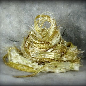 May include: A close-up of a decorative, textured, gold and cream-colored yarn ball. The yarn is made of various ribbons and threads, some with glitter. The yarn is arranged in a loose, layered design.