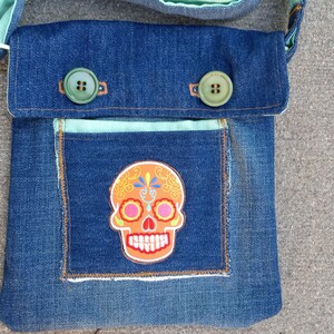 Könnte beinhalten: Eine blaue Jeans-Umhängetasche mit grünem Knopfverschluss und einem bunten Totenkopf-Patch auf der vorderen Tasche.