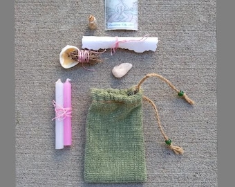 May include: A love spell kit with a green drawstring bag, two pink candles, a rolled up piece of paper tied with pink string, a seashell, a small bottle, a pink stone, and a card with a drawing of a woman with flowers. The text "LOVE SPELL KIT" is written in pink letters on the bottom of the image.