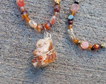 May include: A necklace with a brown and white stone pendant wrapped in silver wire. The necklace is made of brown, orange, and clear beads.