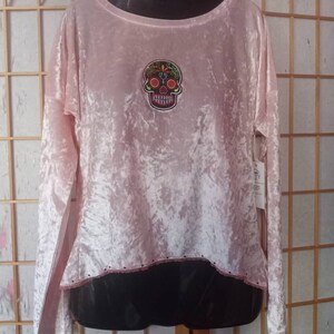 May include: Pink velvet long-sleeved shirt with a black and colourful sugar skull design on the front. The shirt has a scalloped hem.