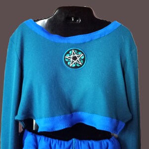May include: A teal long-sleeve crop top featuring a wide blue neckline trim and a matching blue hem. Centered on the chest is a circular patch displaying a white five-pointed star surrounded by stylized green leaves against a black background.