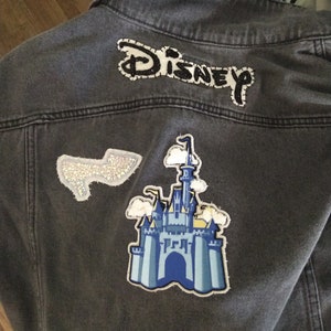 Puede incluir: Una chaqueta vaquera gris con un parche bordado "DISNEY" en blanco y negro, un parche de castillo azul y blanco y un parche plateado brillante.