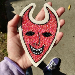 Puede incluir: Parche de tela roja con un dise&ntilde;o de cara de diablo. La cara est&aacute; hecha de lentejuelas rojas y tiene ojos negros y una sonrisa blanca y dentada.