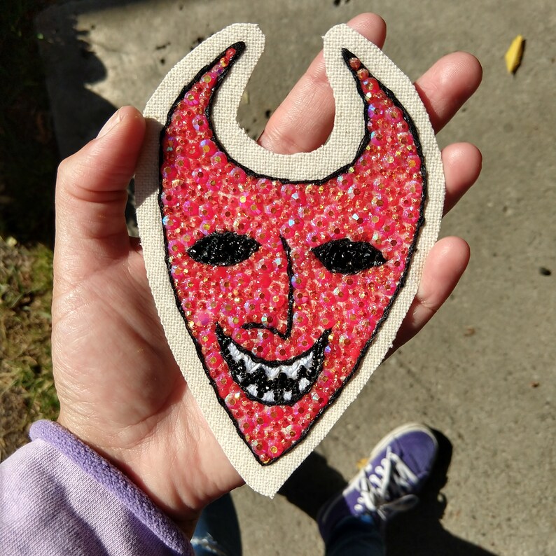 Puede incluir: Parche de tela roja con un dise&ntilde;o de cara de diablo. La cara est&aacute; hecha de lentejuelas rojas y tiene ojos negros y una sonrisa blanca y dentada.