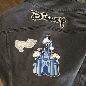Pu&ograve; includere: Una giacca di jeans grigia con una toppa ricamata "DISNEY" in bianco e nero, una toppa di castello blu e bianca e una toppa argentata scintillante.