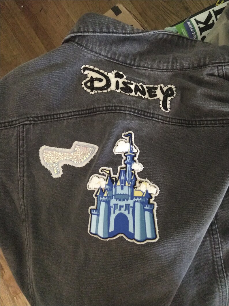 Pu&ograve; includere: Una giacca di jeans grigia con una toppa ricamata "DISNEY" in bianco e nero, una toppa di castello blu e bianca e una toppa argentata scintillante.