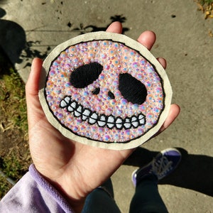 Pu&ograve; includere: Una toppa ricamata rosa e bianca con un design a forma di teschio. Il teschio ha occhi neri e un sorriso bianco. La toppa &egrave; ricoperta di strass iridescenti.