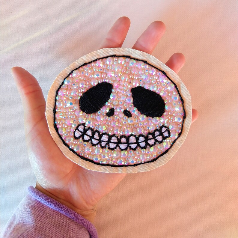 Pu&ograve; includere: Un cerotto di tessuto bianco con un design di teschio con perline rosa e iridescenti. Il teschio ha occhi neri e un sorriso cucito.