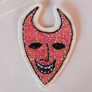 Puede incluir: Una m&aacute;scara de diablo roja con ojos negros y una sonrisa de dientes, hecha de lentejuelas y cuentas iridiscentes sobre un fondo de tela blanca.