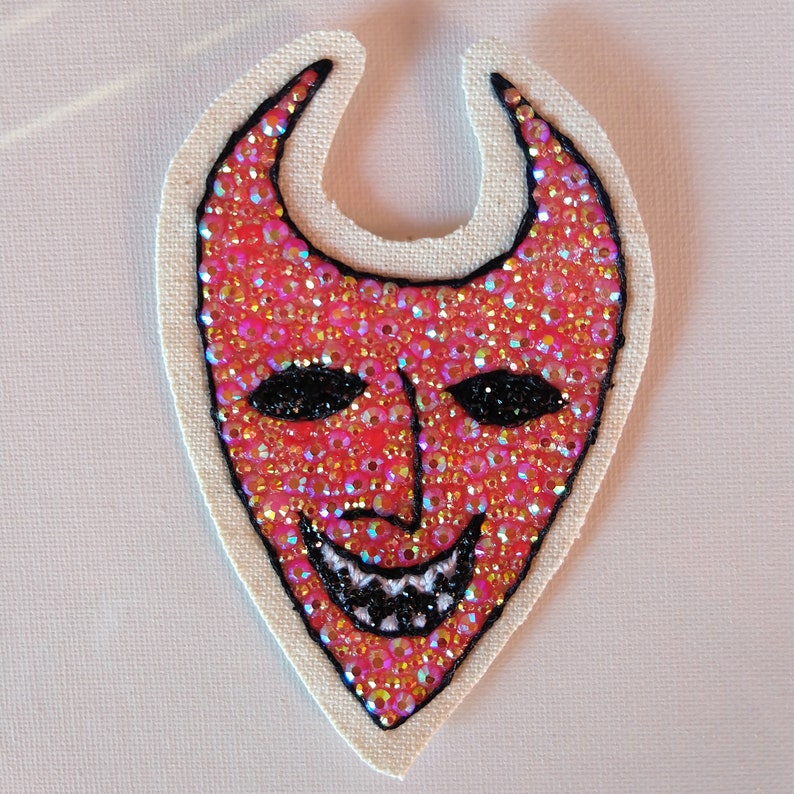 Puede incluir: Una m&aacute;scara de diablo roja con ojos negros y una sonrisa de dientes, hecha de lentejuelas y cuentas iridiscentes sobre un fondo de tela blanca.