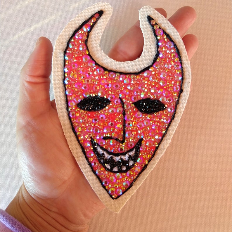 Puede incluir: Un parche de tela rojo y blanco con un dise&ntilde;o de cara de diablo. La cara est&aacute; hecha de pedrer&iacute;a iridiscente y tiene una sonrisa cosida negra.