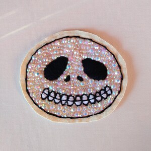 May include: A white fabric patch with a black embroidered skull design. The skull has a large smile with teeth and is covered in iridescent pink and white beads.