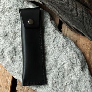 May include: A black leather pencil case with a stitched edge and a metal button closure.