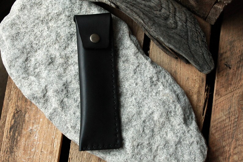 May include: A black leather pencil case with a stitched edge and a metal button closure.