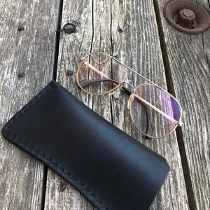 May include: A black leather glasses case with stitching detail, lying on a weathered wooden surface. A pair of silver-rimmed glasses with clear lenses is resting on the case.