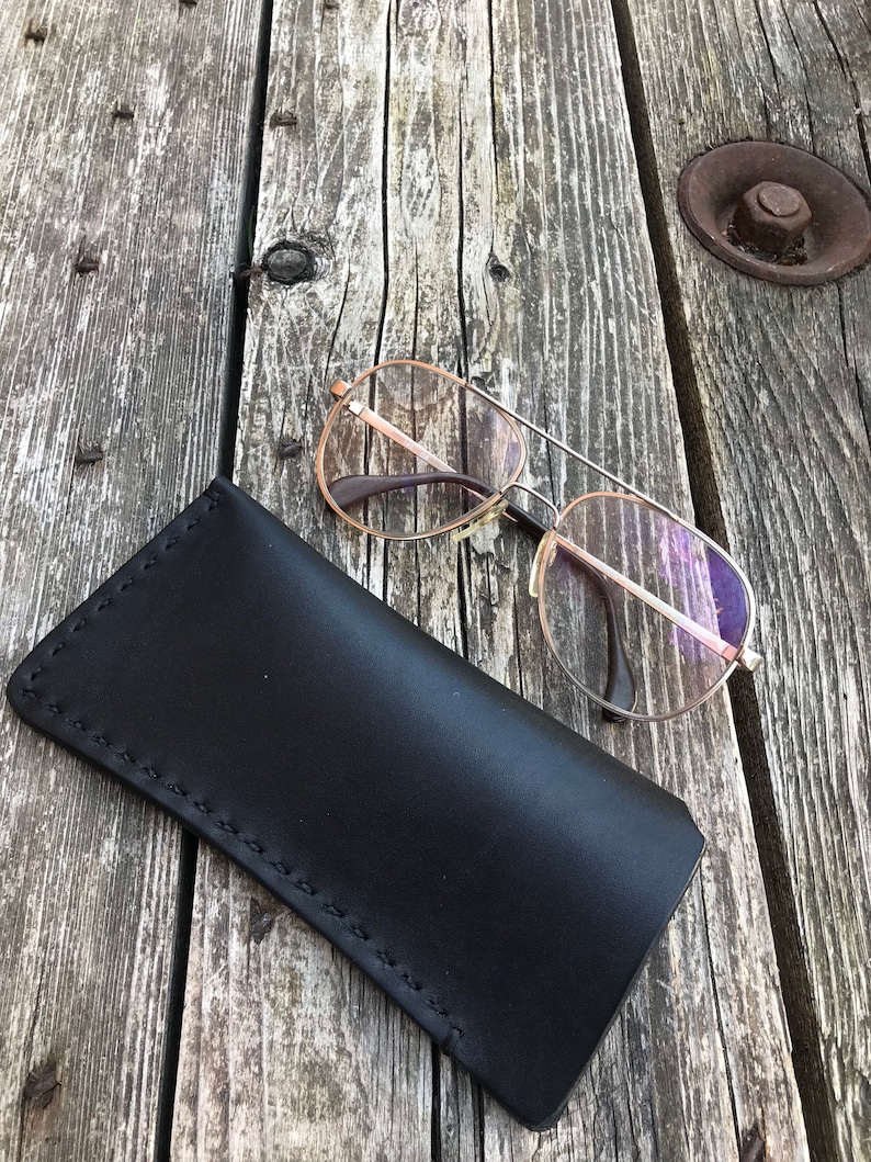 May include: A black leather glasses case with stitching detail, lying on a weathered wooden surface. A pair of silver-rimmed glasses with clear lenses is resting on the case.