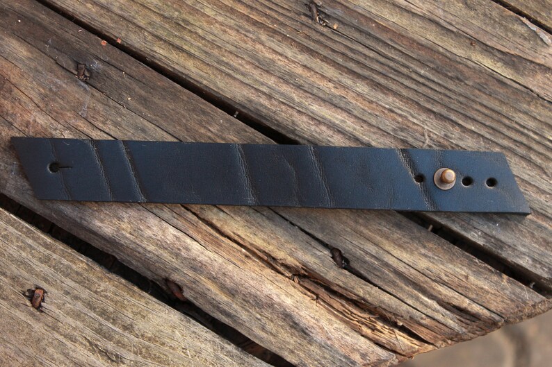 May include: A black leather bracelet with a brass buckle and three holes. The bracelet has a textured surface.