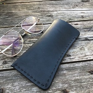 May include: A black leather glasses case with a stitched edge. The case is lying on a wooden surface.