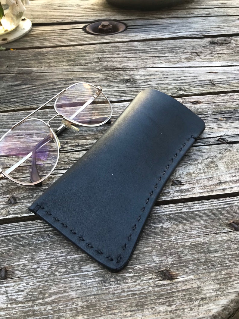 May include: A black leather glasses case with a stitched edge. The case is lying on a wooden surface.