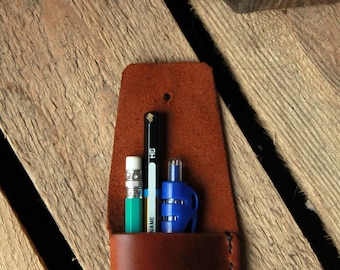 May include: A brown leather pencil case with a stitched edge and a metal button closure. Three writing utensils are inside: a black pencil, a green pencil, and a blue pen.