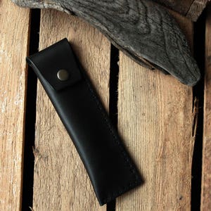 May include: A black leather pencil case with a stitched edge and a metal button closure.