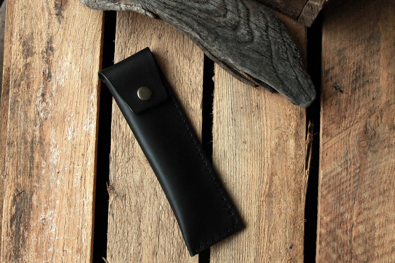 May include: A black leather pencil case with a stitched edge and a metal button closure.