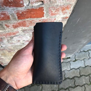 May include: A black leather case with stitching around the edges. The case is held in a hand.