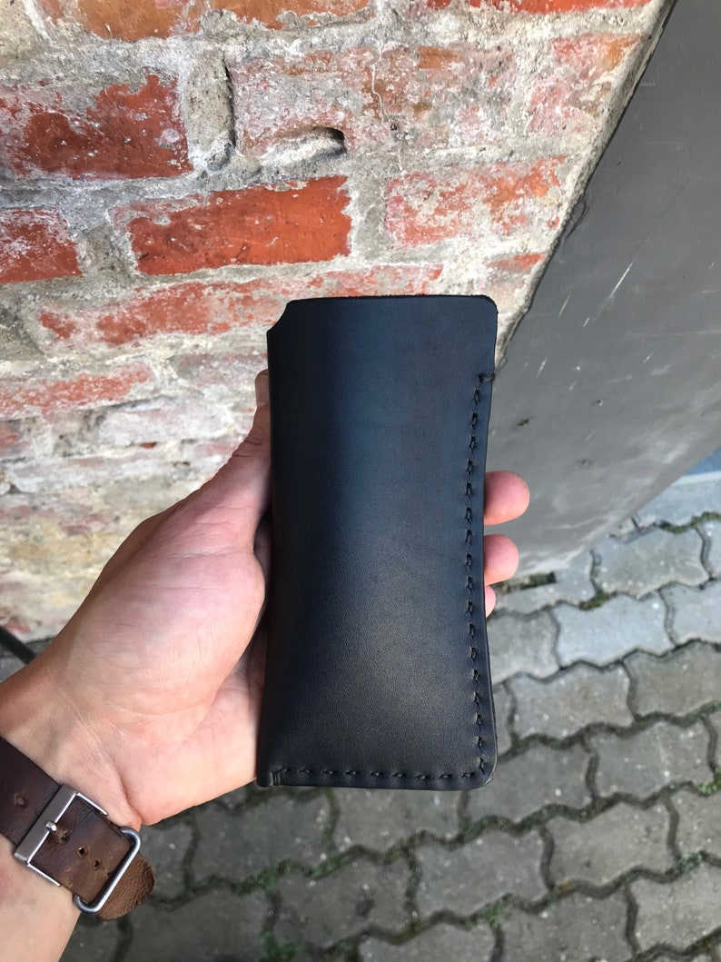 May include: A black leather case with stitching around the edges. The case is held in a hand.