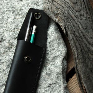 May include: A black leather pencil case with a snap closure, holding a black pen and a green pencil.