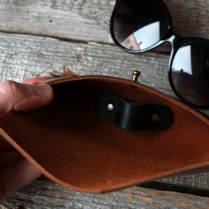 May include: A brown leather sunglasses case with a black leather strap and a brass clasp. The case is open and the inside is visible.