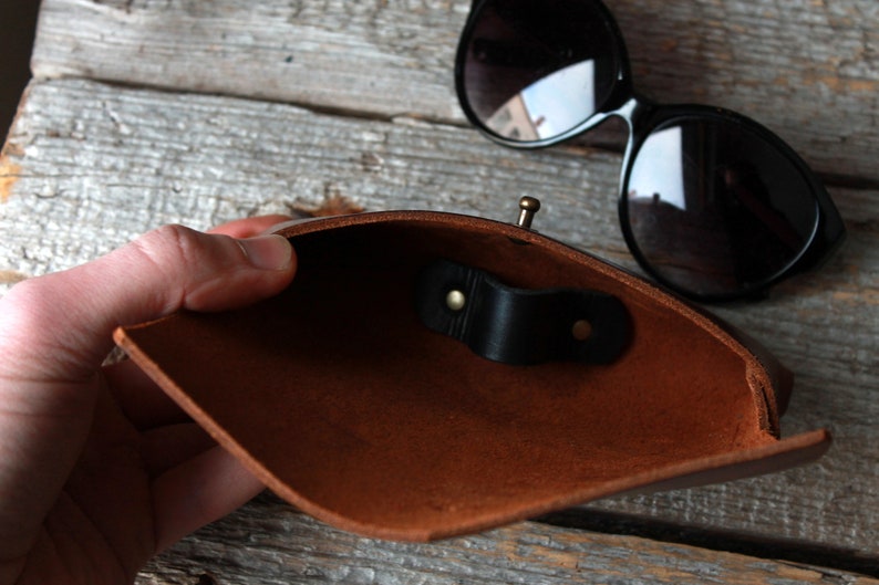 May include: A brown leather sunglasses case with a black leather strap and a brass clasp. The case is open and the inside is visible.