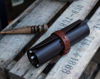 May include: A rolled-up brown leather case with a brown leather strap and a brass buckle. The case is sitting on a wooden surface with the text "8889 1717 TEL. - TURIL - 000" printed on it.