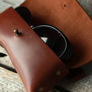 May include: A brown leather sunglasses case with a brass button closure and two brass rivets. The case is open and a pair of black sunglasses are inside.