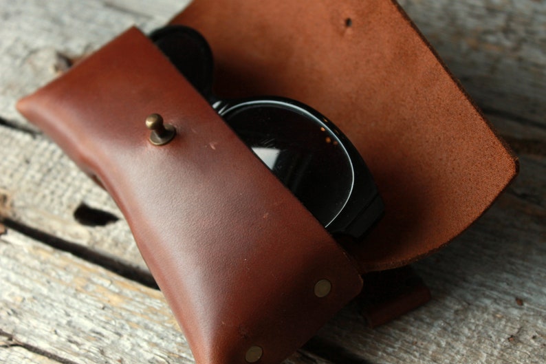 May include: A brown leather sunglasses case with a brass button closure and two brass rivets. The case is open and a pair of black sunglasses are inside.