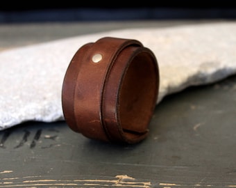 May include: A brown leather cuff bracelet with a silver rivet. The bracelet is made of multiple layers of leather and is rolled up.