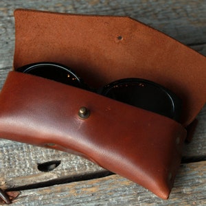 May include: A brown leather case for sunglasses with a snap closure. The case is open and two pairs of sunglasses are inside.