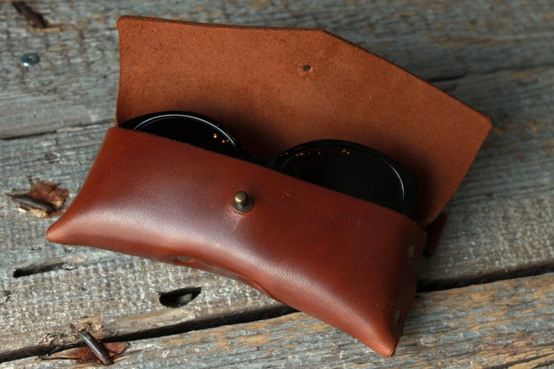 May include: A brown leather case for sunglasses with a snap closure. The case is open and two pairs of sunglasses are inside.